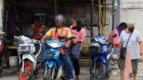 A woman hops on the back of a motorcycle taxi. Drivers sit in stands waiting for passengers to show up